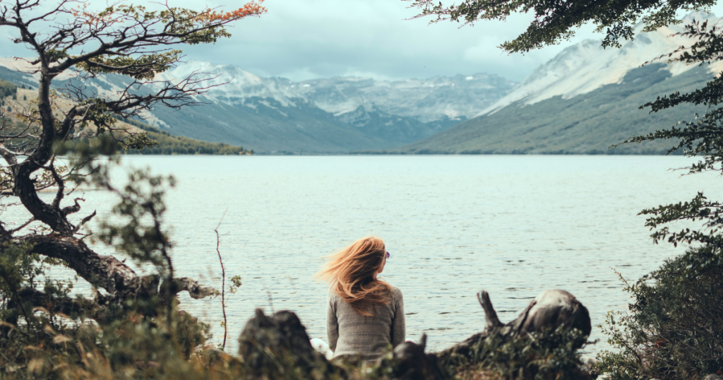 Person sitting quietly by a peaceful lake surrounded by mountains and trees, symbolizing rest and recovery from burnout.