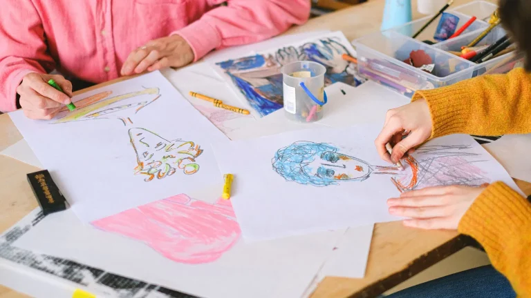 Two people drawing with crayons and oil pastels at a shared table covered in colourful artwork, representing the Second Spring art therapy group for women in perimenopause and menopause in Ottawa.